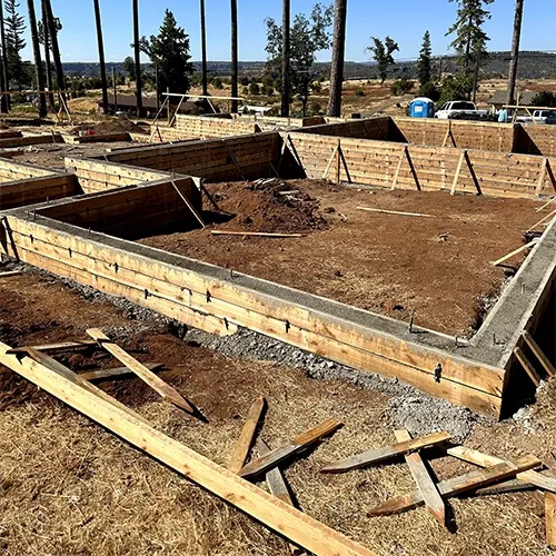 A construction site shows a rectangular concrete foundation with wooden formwork. Dirt fills the center, and wooden braces support the forms. Scattered lumber lies in the foreground. Tall trees, open land, and blue portable toilets are visible in the background.