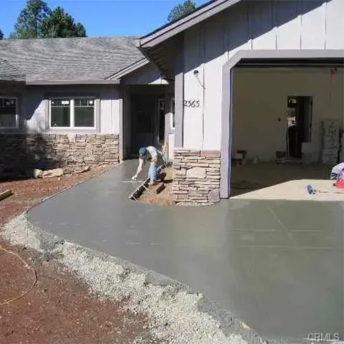 A person in work clothes smooths freshly poured concrete on a driveway leading to a house with gray siding and stone accents. The garage door is open, and some tools and supplies are visible inside. Another person stands inside the garage.